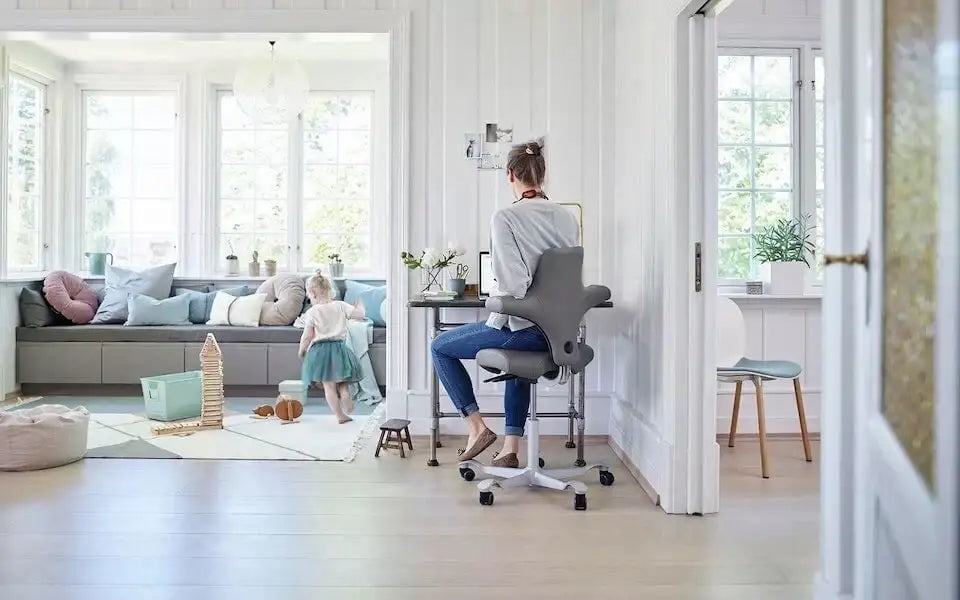 osoba siedząca na ergonomicznym krześle biurowym w jasnym, przestronnym wnętrzu z dzieckiem bawiącym się na dywanie