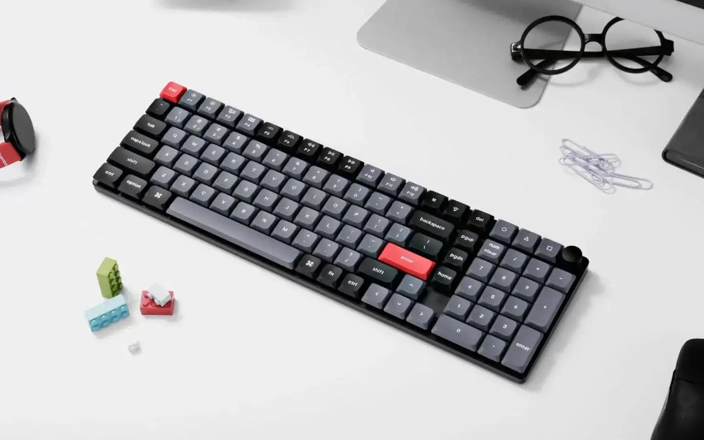 Mechaniczna klawiatura z szaro-czarnymi klawiszami i czerwonymi enter i ESC na białym biurku z okularami i zegarkiem