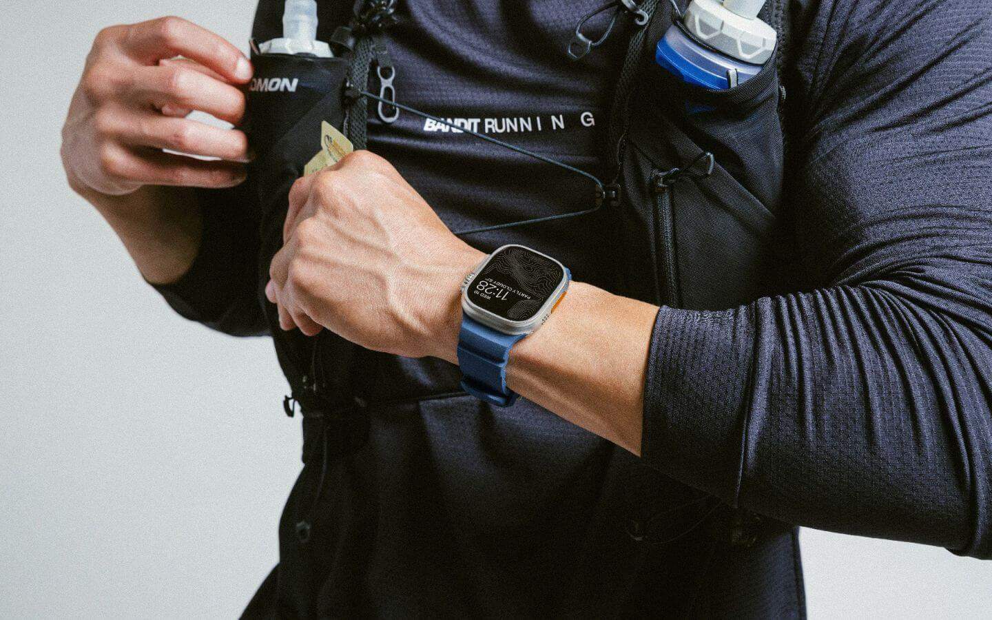 Biegacz w czarnej odzieży sportowej z zegarkiem sportowym i bidonem Salomon w plecaku do biegania