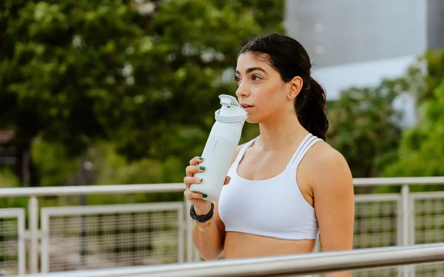 Kobieta w białym topie sportowym pije wodę z białej butelki na zewnątrz przy poręczy z zielonym tłem