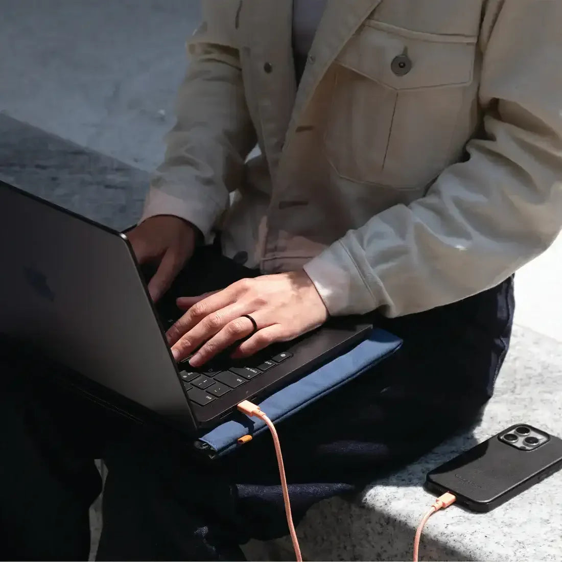 Osoba pracująca na laptopie Apple z beżową kurtką, laptop na poduszce, ładowany kablem apricot obok smartfona na kamiennej ławce