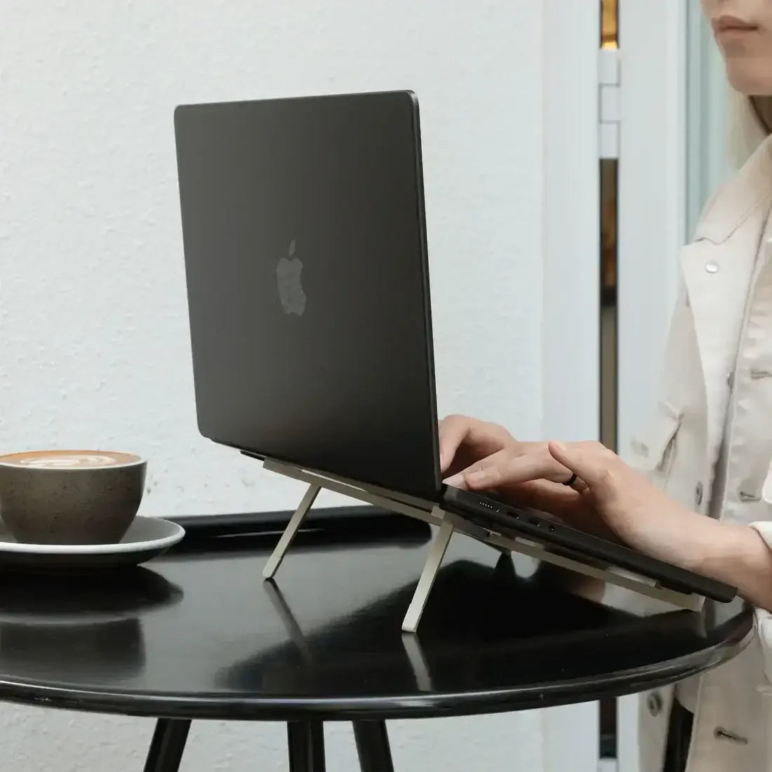 Czarny laptop na składanym stojaku na stole obok filiżanki kawy, osoba pisząca na klawiaturze