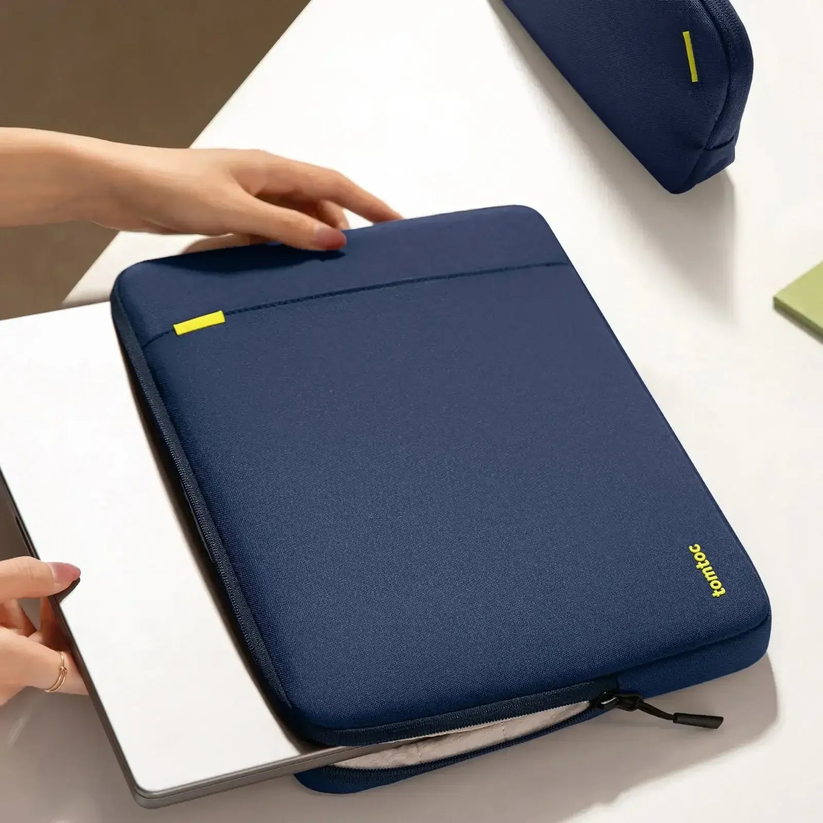 Granatowe etui na laptopa na biurku, ręka wkłada komputer, minimalistyczny design, akcesoria biurowe