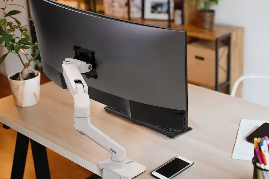 Biurko home office z zakrzywionym monitorem na regulowanym uchwycie, smartfonem i rośliną.