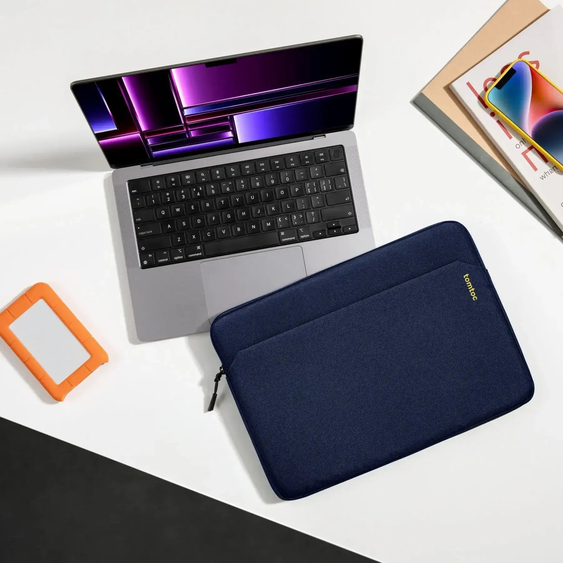 Laptop na biurku, niebieskie etui ochronne na laptopa, dysk zewnętrzny, smartfon i czasopisma. Idealny zestaw do pracy zdalnej i home office.