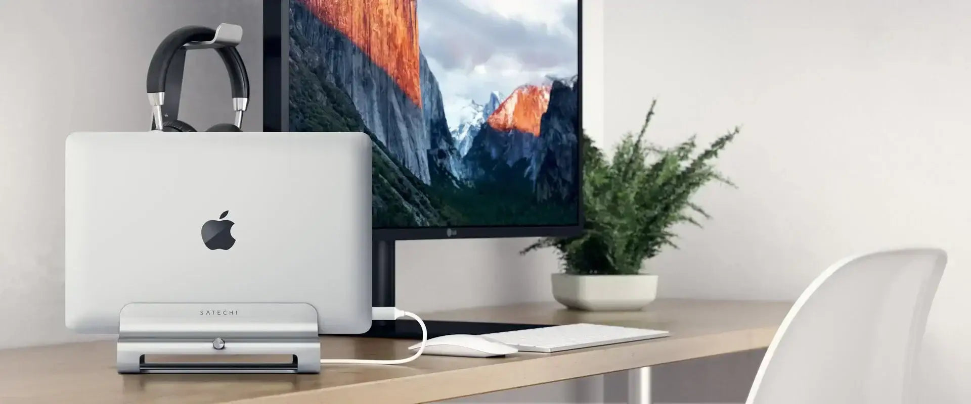 Laptop Apple na srebrnym stojaku Satechi pionowo obok monitora na minimalistycznym biurku z rośliną