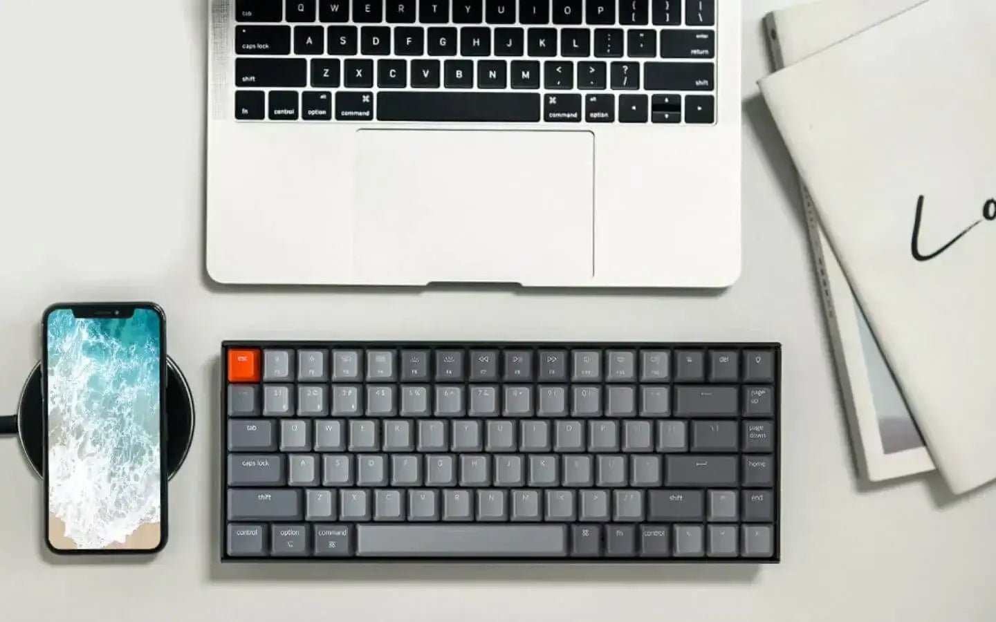 Biurko z laptopem, czarno-szarą klawiaturą mechaniczną i smartfonem na ładowarce bezprzewodowej