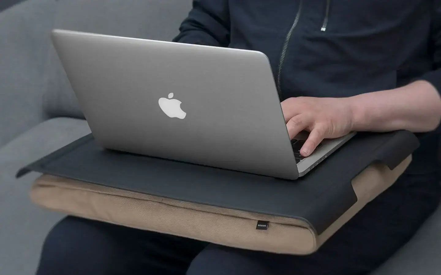 Osoba pracująca na laptopie Apple na poduszce na kolanach, siedząc na kanapie
