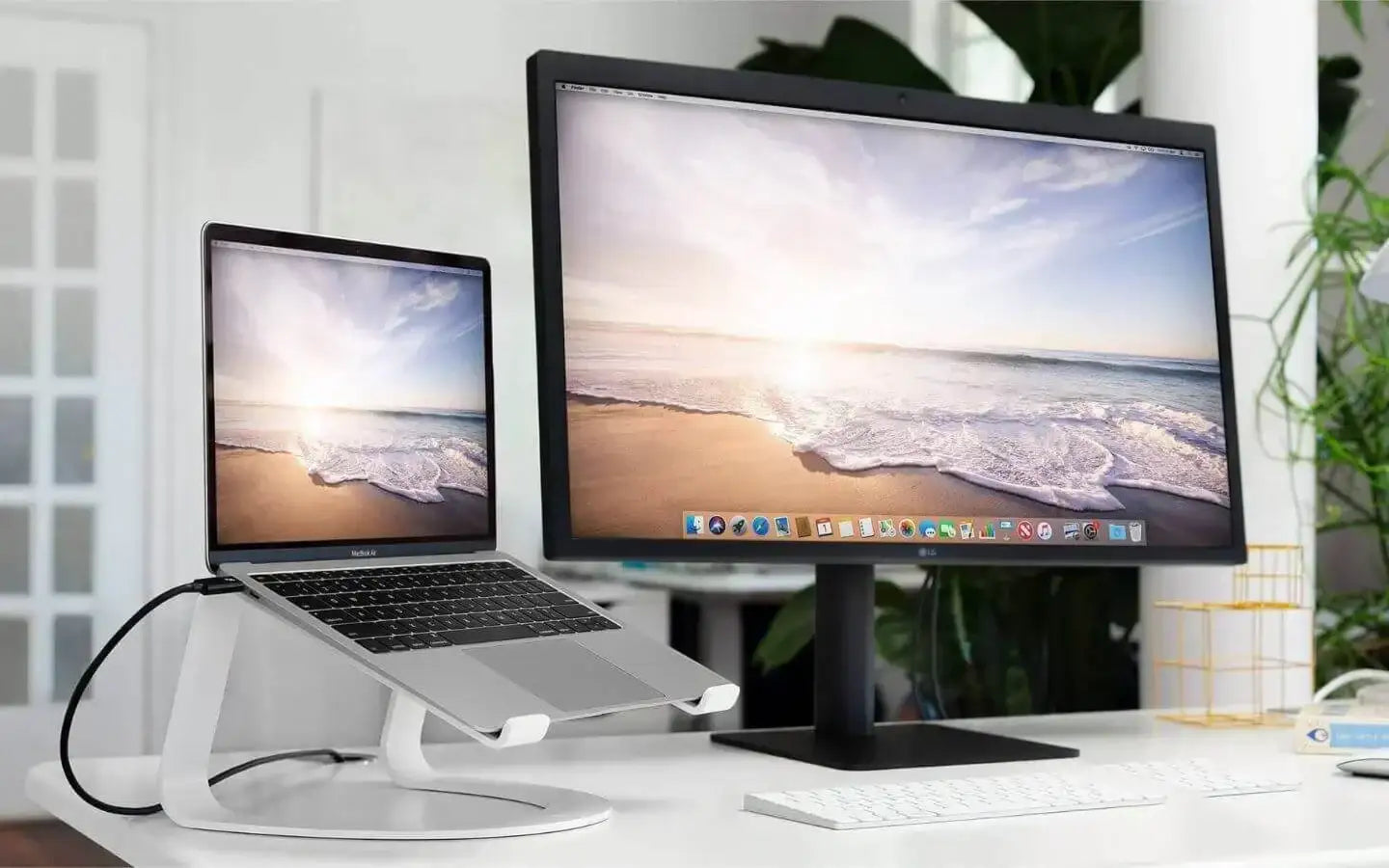 Laptop MacBook na białym stojaku obok monitora z obrazem plaży i oceanu na białym biurku