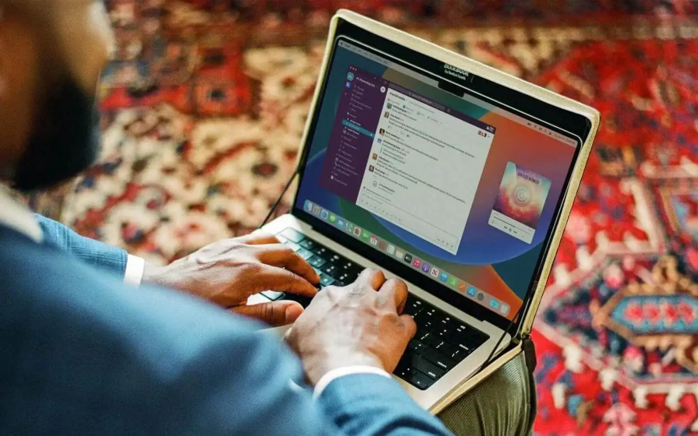 Mężczyzna korzystający z laptopa MacBook z aplikacją Slack na tle kolorowego dywanu