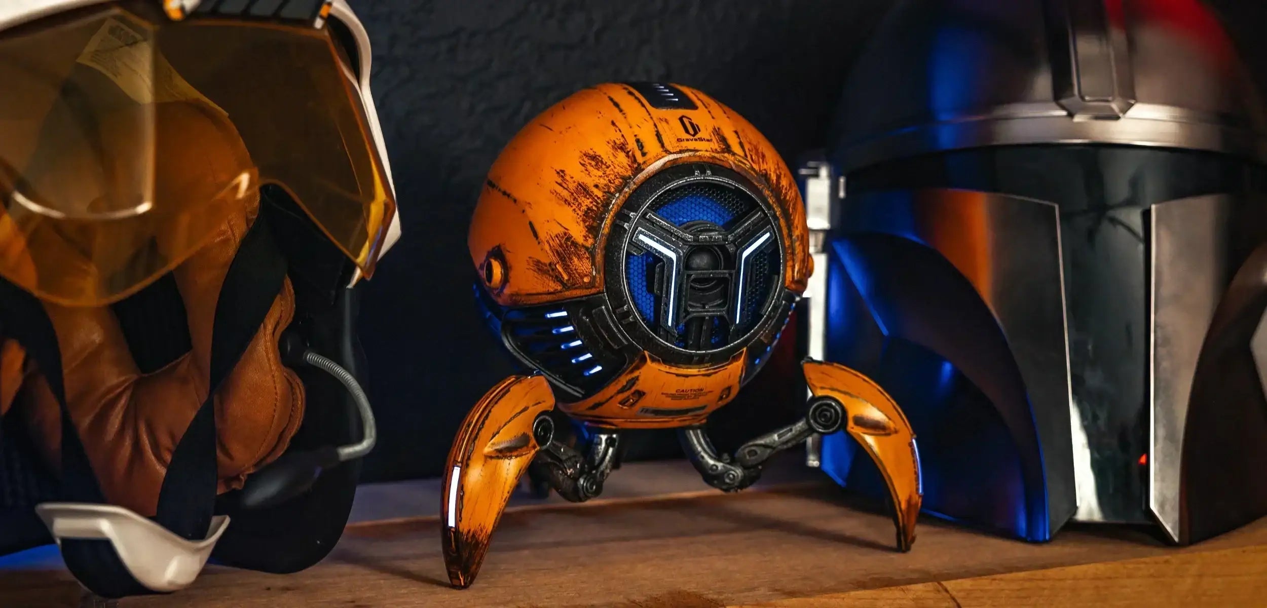 Futurystyczny pomarańczowy robot z trzema ruchomymi nogami między kaskiem lotniczym i hełmem Mandalorianina na drewnianej powierzchni