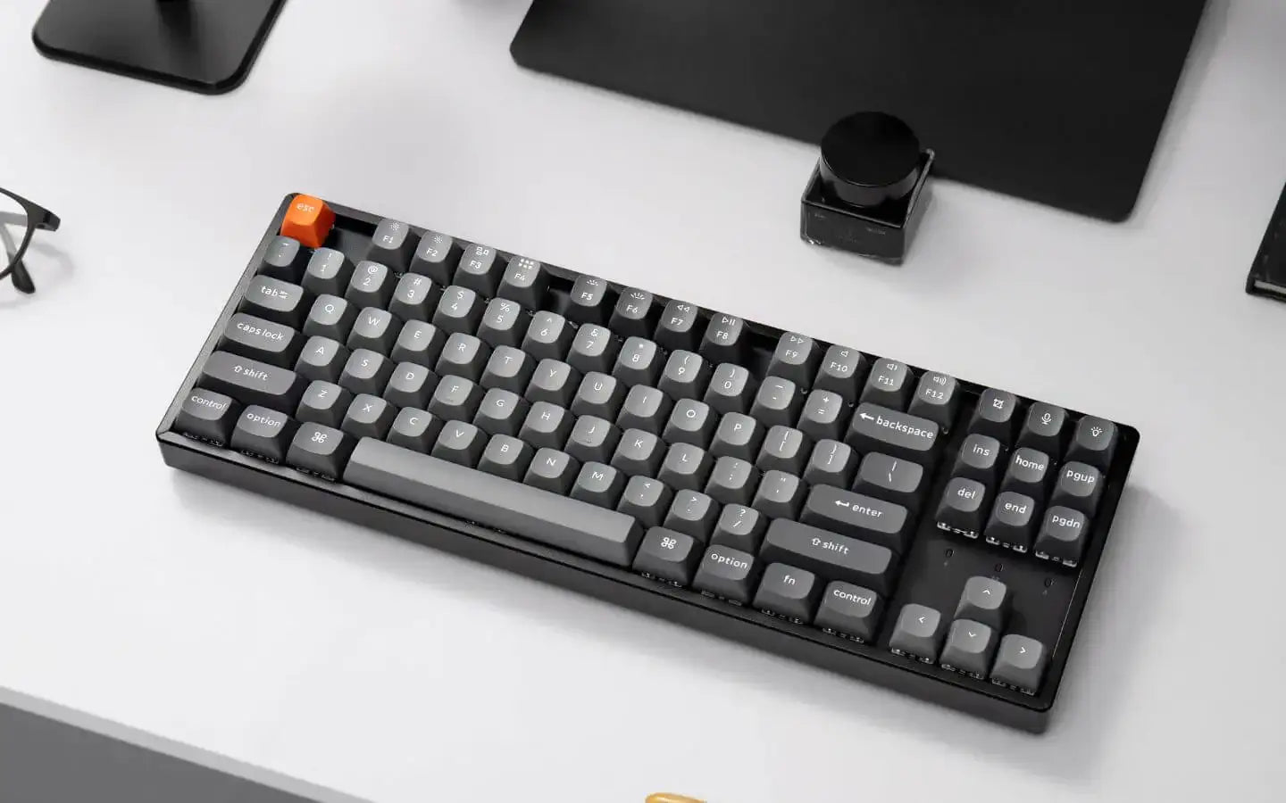 Czarna mechaniczna klawiatura z szarymi klawiszami i pomarańczowym klawiszem ESC na białym biurku