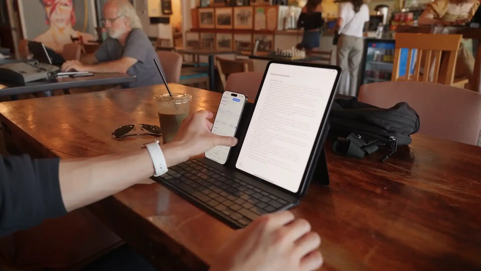 Osoba pracująca na tablecie z klawiaturą i smartfonem, chłodna kawa na drewnianym stole w kawiarni