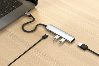 Srebrny adapter USB-C 4w1 podłączony do laptopa na drewnianym biurku, widoczne złącza USB i HDMI
