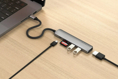 Srebrny adapter USB-C z wieloma portami podłączony do laptopa na biurku, widoczne pendrive'y i karta SD.