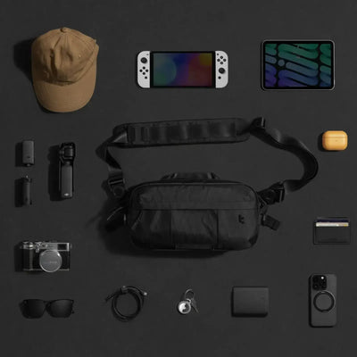 Czarna torba na ramię oraz akcesoria EDC: czapka, kamera, konsola, tablet, okulary, portfel, powerbank.